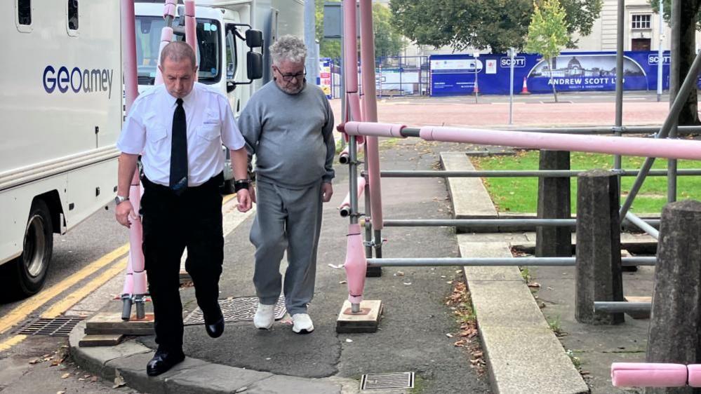 Anthonio Villafane is wearing a grey tracksuit with white trainers. He has grey/white curly hair and black glasses. He looks down as he is led by a Geoamey court officer who is wearing a white shirt, black trousers, black boots and a black tie. Anthony Villafane is handcuffed to the court officer by the wrist. 