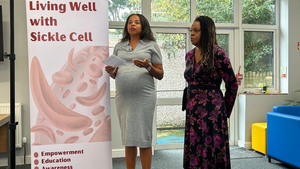 Rochelle Raheem in a grey dress standing next to Catherine James-Oliver in a black and purple floral dress with a sickle cell banner to their right.