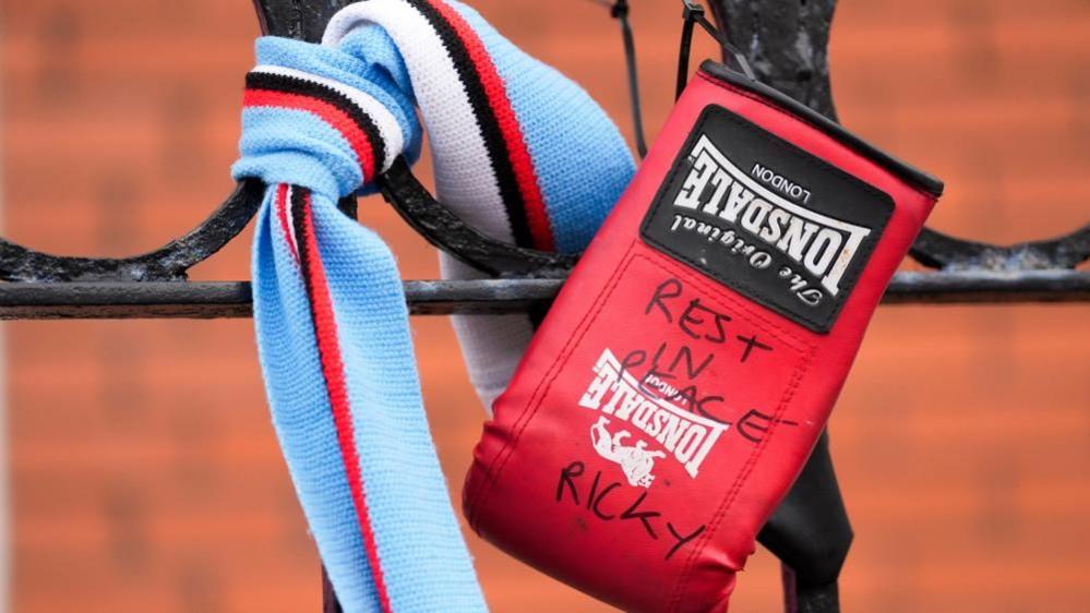 A red boxing glove hangs from a fence with the words "Rest in peace Ricky" written on it. A blue, white, red and black striped scarf is tied on the fence next to it. 