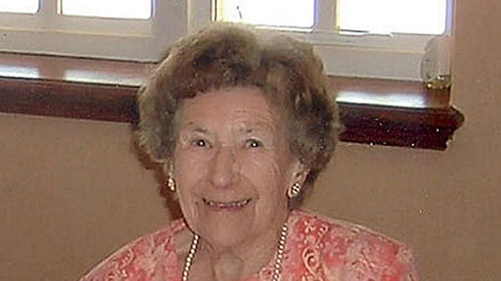 Una Crown smiling at the camera while sitting in front of a beige-coloured wall and window.  She is wearing a pink patterned dress with a pearl necklace and shiny earrings.