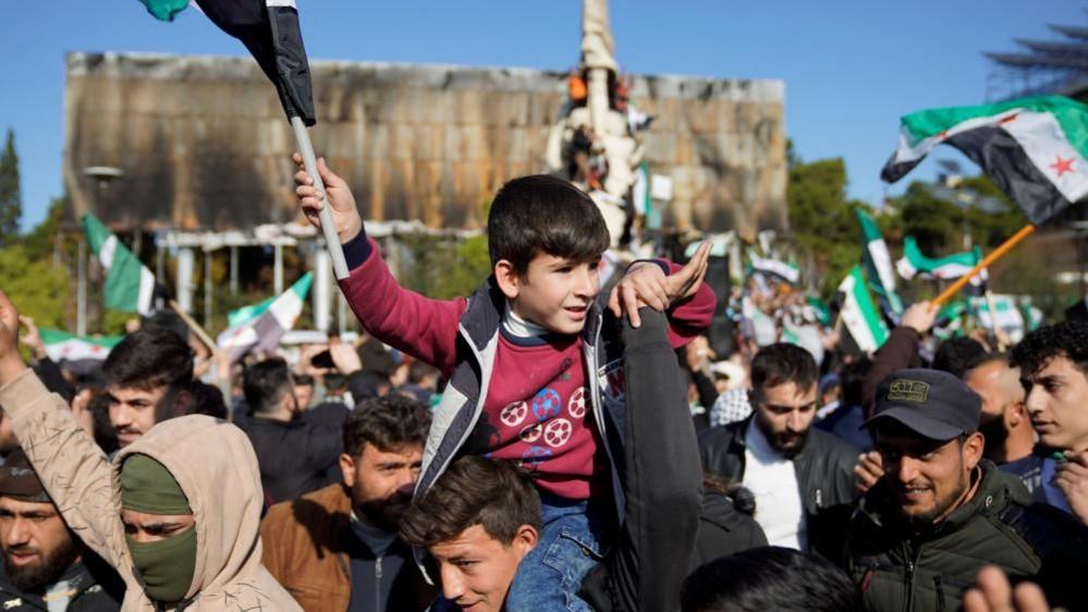 A boy sits on the shoulders of an adult man and waves a Syrian flag, many men in the crowd are also waving Syrian flags in celebration.