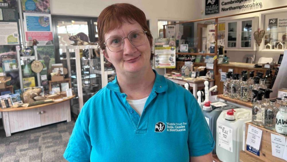 Julia French, wearing a blue polo shirt, with short red hair, wearing glasses, looking at the camera, in a shop, with items to buy around her. 