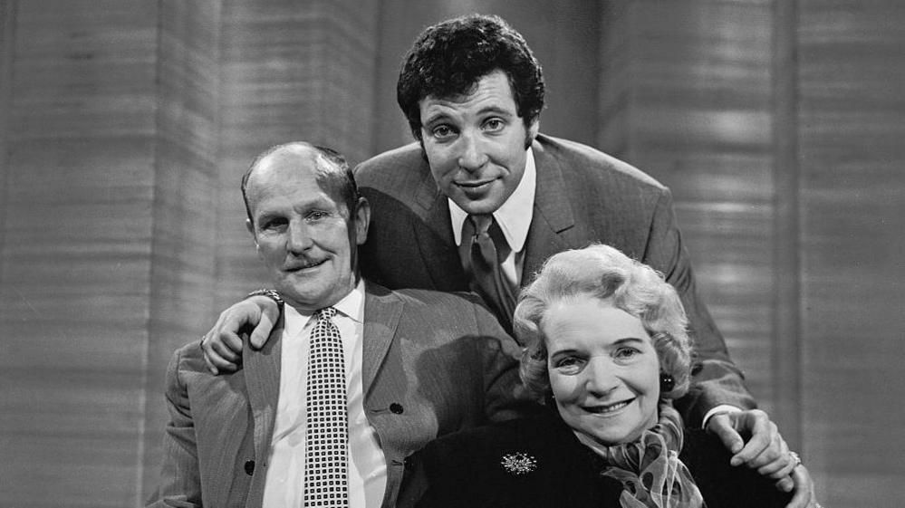 Black and white photo. Tom Jones stands behind his parents, who are sitting. He has his arms around them and they all smile at the camera. They are all dressed in smart clothes, Tom Jones and his father are in suits and his mother is wearing a black coat with a broach and scarf.