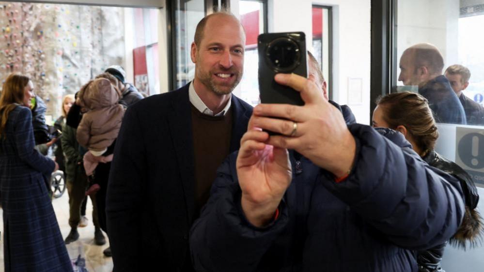 Princi buzëqesh ndërsa shikon në një telefon celular të cilin një person me xhaketë blu e mban pranë tij.