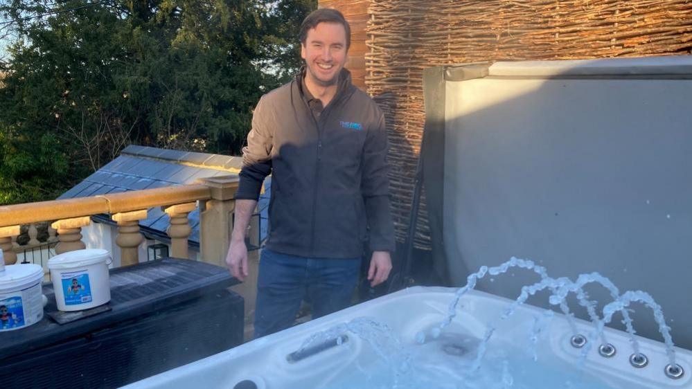 James Symons standing behind of a hot tub with jets of water shooting in the air. He is wearing a brown jumper and smiling at the camera