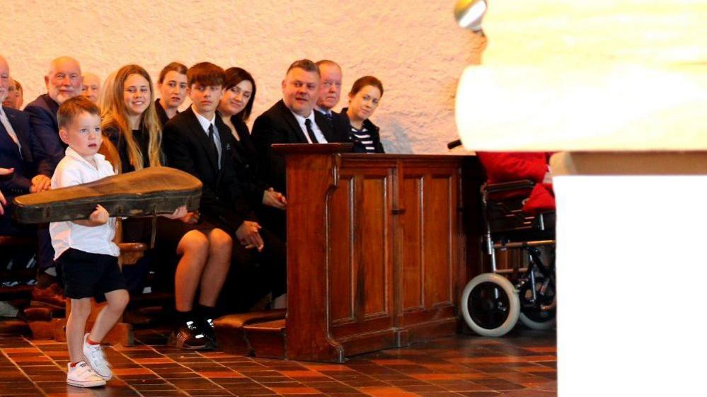 A young boy carrying a fiddle up a tiled floor at a funeral