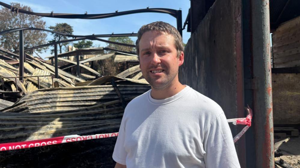 Matthew stood in front of the remains of one of the barns. He's wearing a white tshirt and is looking into the camera with a bit of a grimace. Behind him, is a huge pile of twisted steel beams and collapsed corrugated steel panels. The metal is blackened and covered in dust. The sky is bright blue and it's clearly sunny. Behind Matthew there's a red and white tape blocking access to the collapsed barn, reading 'Do not cross.'