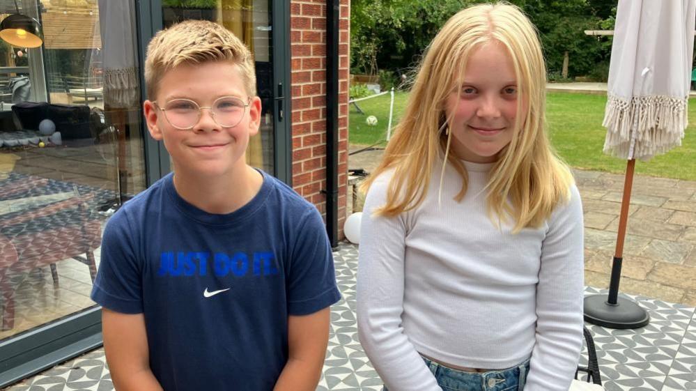 A boy and girl look into the camera, standing in a garden.