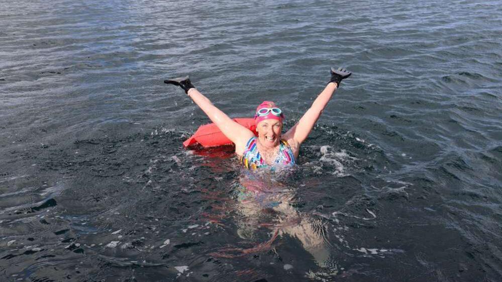 Jules is in the sea wearing a swimming costume, a swimming cap, black gloves and goggles on top of her head, as well as a flotation device. She is holding her hands up in the air and smiling at the camera.