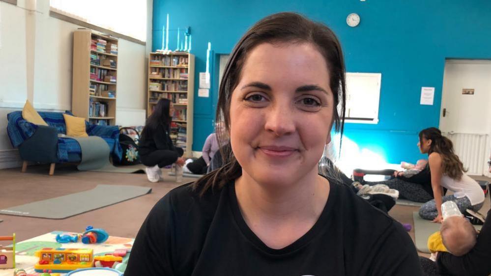 A woman is sitting on a mat with baby toys and mums and babies in the background. She is wearing a black t-shirt, has long dark hair and is smiling at the camera