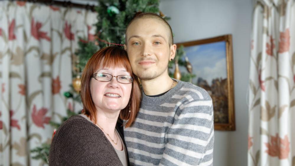 Robin Freeman, who was diagnosed with testicular cancer in his early 20s, has short hair and is wearing a grey and white striped top. He is hugging his mum, who has auburn hair and is wearing a brown jersey. They are pictured in a room which features floral patterned curtains and pictures in the background.