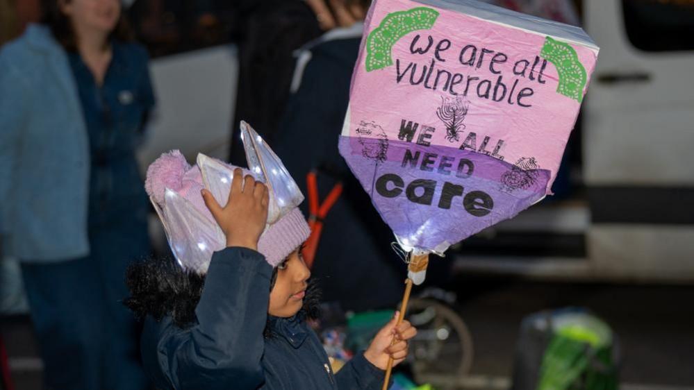 A young girl at a protest holds up a placard that reads 'We are all vulnerable, we all need care'. She is wearing a crown-shaped piece of headgear which has small lights in it