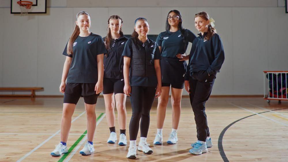 A group of five girls standing in a school sports hall. There are basketball hoops and a trolley in the background. Each girl is wearing a different outfit with different parts of the kit, including black leggings, a black gilet, a navy blue half-zip jumper and black jogging pants.