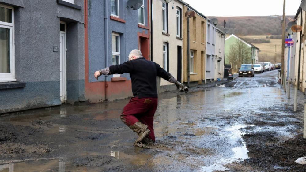 A man with grey hair, a dark top and red trousers walks to his home through deep mud covering a street in the aftermath of Storm Bert