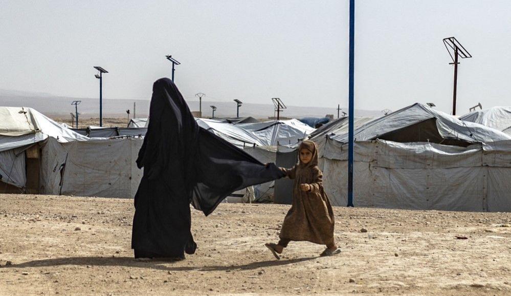 A small child in a brown dress tugs on the back of a woman's niqab as they walk past white tents on sandy ground