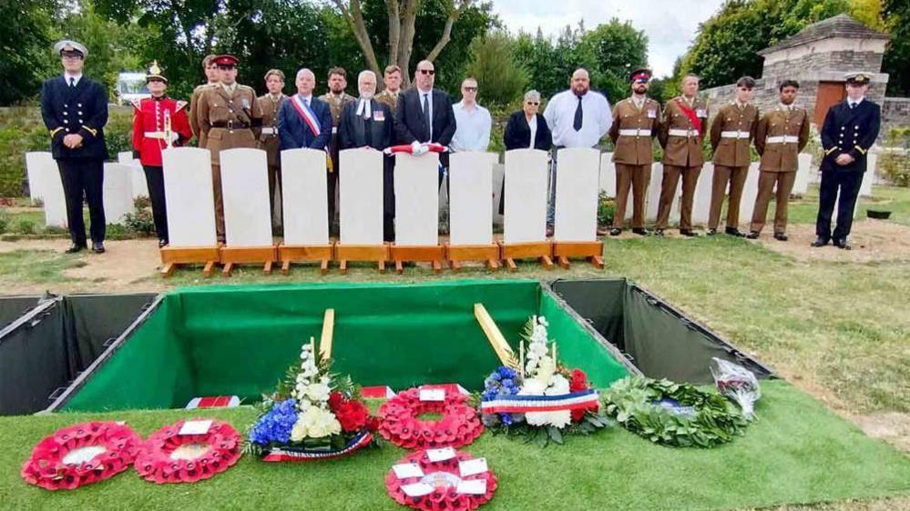 A burial ceremony with soldiers standing in front of the spaces where the coffins are going to be lowered into, and various wreaths in the foreground.