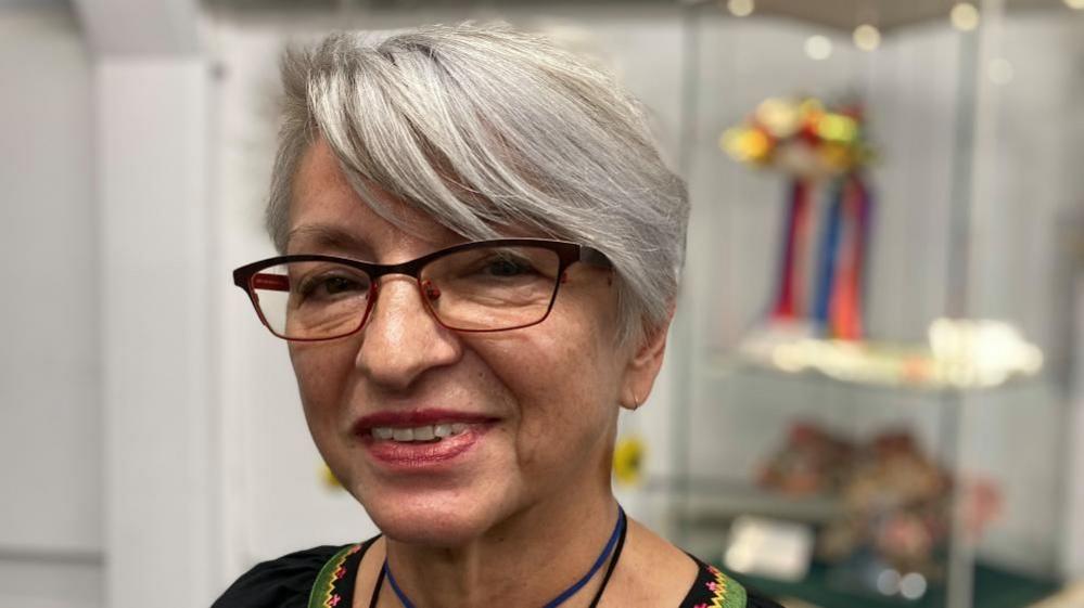 A lady, Raissa, 70, looks directly at the camera. She wears burgundy glasses and her grey hair is cut in a bob hair style.