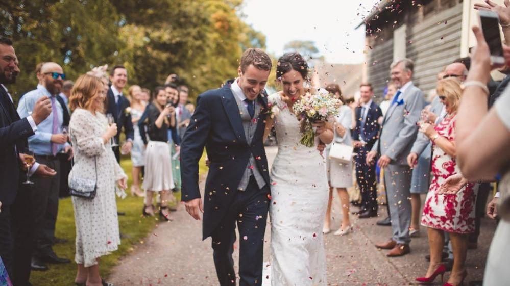 A newly married couple walk through a tunnel of people, who are throwing confetti at them. The bride is on the right dressed in white and carrying a boquet. The groom is on the left in a blue suit with a grey waistcoat. 