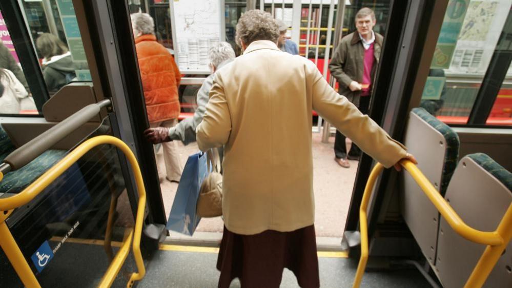 An elderly lady in a light mustard coat steps off a bus. She holds on to yellow rails with her right arm. A bus stop with a number of people can be seen in the background.