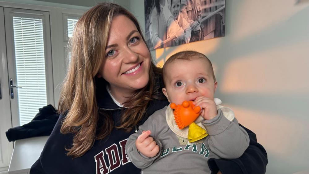 Charlie Hawksworth holding Roman, who has a toy in his mouth and a grey top on. Charlie is smiling, with her head slightly tilted to the left. They are are in a room with a picture on the wall and doors behind her. 