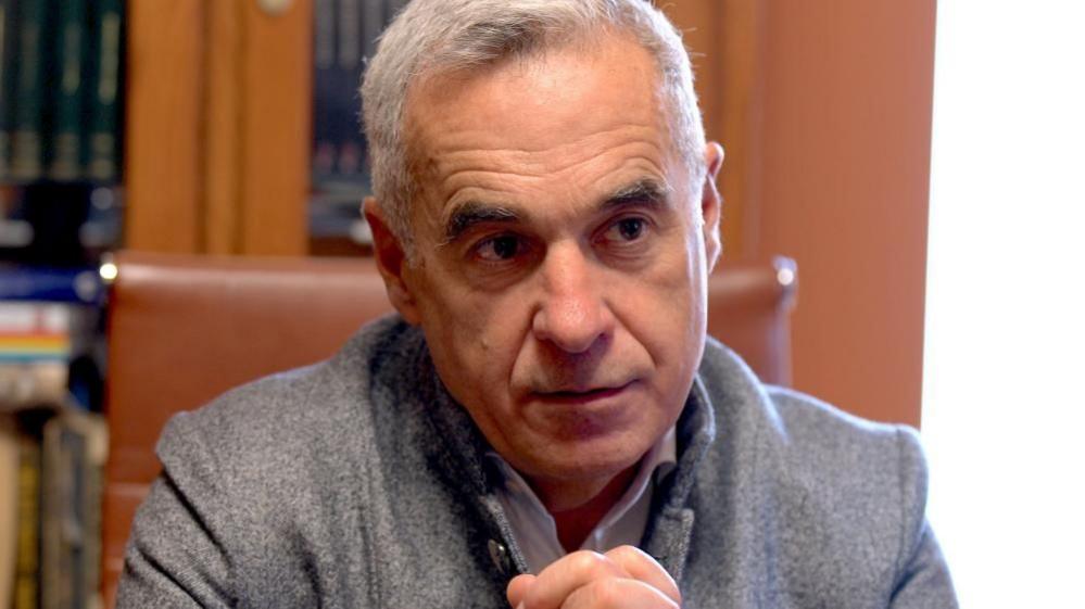 Georgescu, a man with grey hair and wearing a grey suit, sits at a desk, his hands clasped in front of him