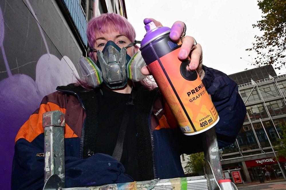 A woman is holding spray paint cans while standing on a ladder. She is wearing a large oversized multi-colour coat and a large building is visible behind her. 