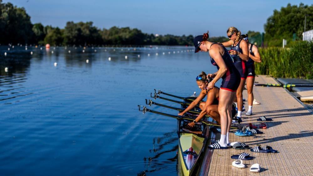 Four women in blue and red outfits stand on the bank with a canoe in the water