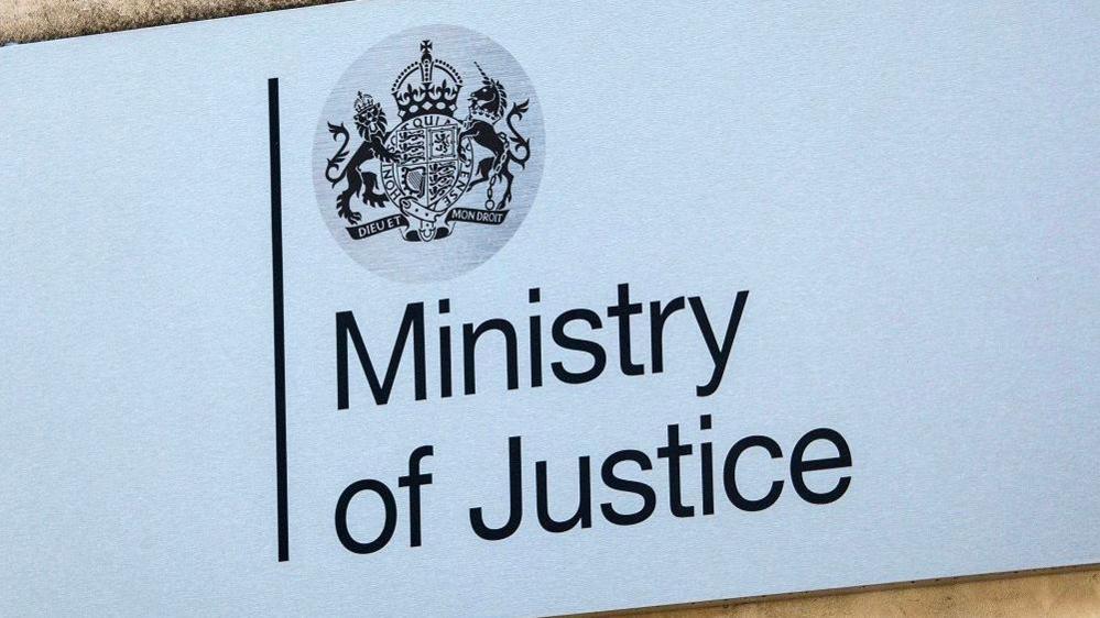 A grey Ministry of Justice sign with the words typed in black letters. The Government. The coat of arms of the United Kingdom is above, featuring a lion, unicorn and crown above some Latin words.