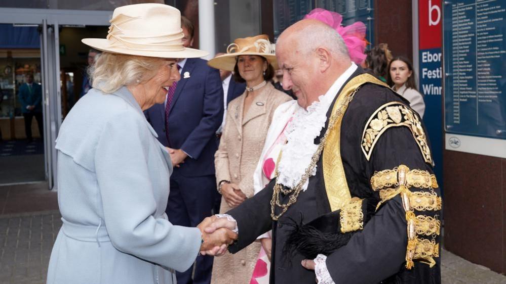 Queen Camilla greets Lord Mayor of York, Councillor Martin Rowley BEM. She wears a light coloured hat and a pale blue trench coat. The mayor is dressed in official robes of black and gold, with a ceremonial chain.