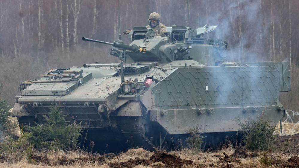 A Warrior tracked armoured vehicle on rough grassland in front of trees.