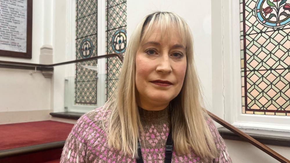 Melanie Holland has long blond hair, and is wearing a pink and brown jersey. She is standing on the red-carpeted staircase at Great Yarmouth Town Hall. There are stained glass windows behind her.