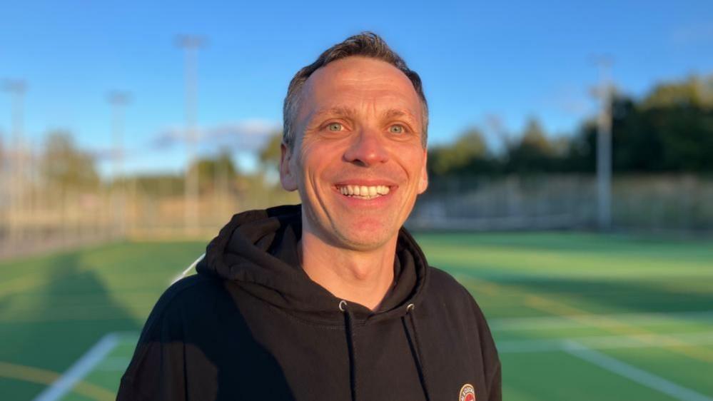 A middle-aged white man with brown hair is standing on an artificial football pitch, smiling broadly in the setting sun. He is wearing a black hooded top with a football emblem on the chest.