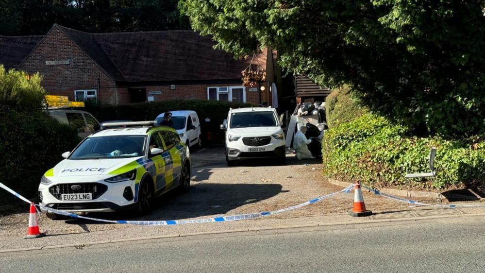 Four vehicles outside a property in Brenwood, showing a forensic officer in a white hazmat suit. There is also police tape and cones. 