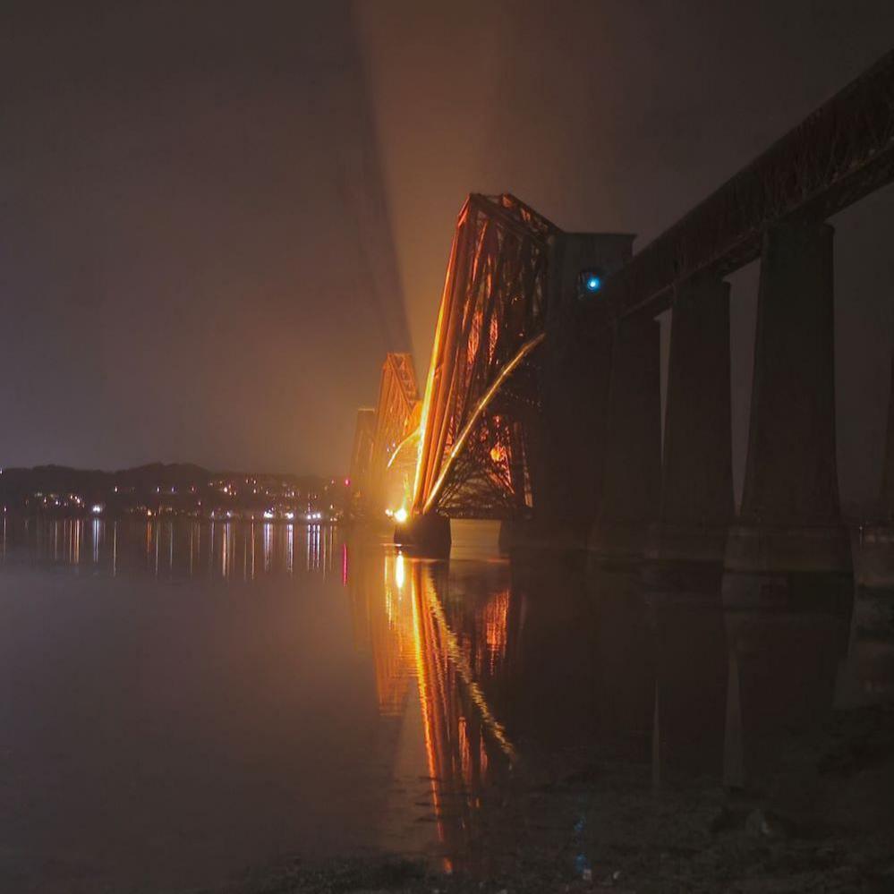 The Forth Bridge at night with some mist and an orange light highlighting the structure.