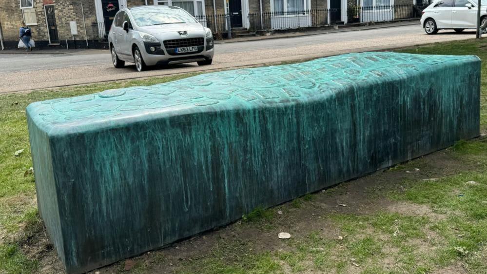 A copper coloured rectangle, with relief designs on the top. It forms a public bench on the edge of St George's Park. Houses and cars are parked in the backdrop.