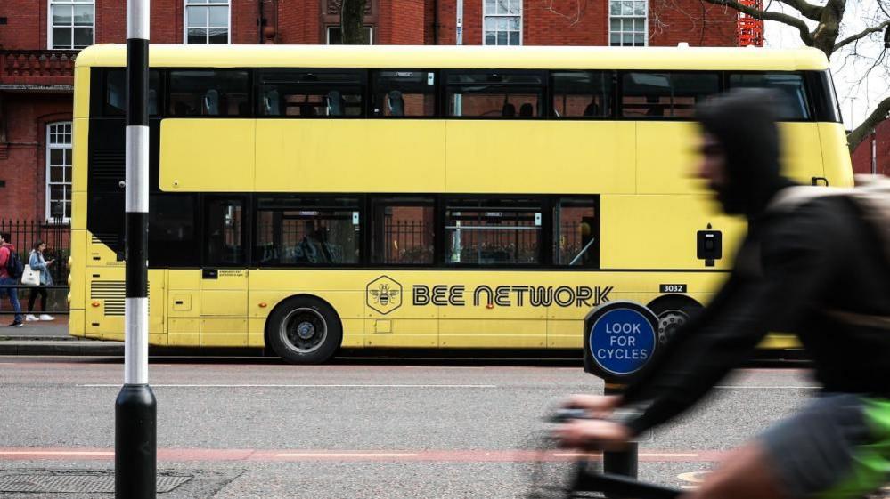 A yellow double decker bus with 'Bee Network' written on the side is stopped at a bus stop. A person rides his bike in the foreground. 