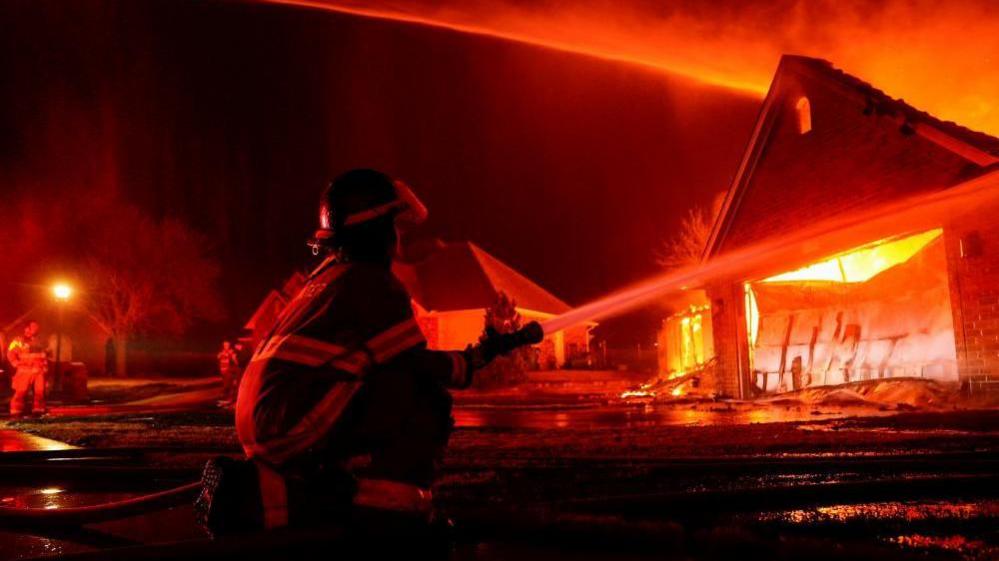 A firefighter crouching down and hosing a garage that is full of flames.