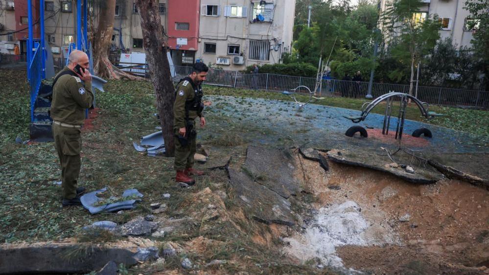 Two Israeli military personnel wearing green combat gear examine a crater left by a Houthi missile attack in Tel Aviv. In the background are swings and other playground apparatus.