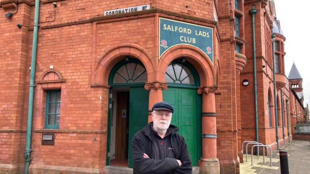 A middle-aged man with a white beard and a cap stands with his arms folded in front of Salford Lads Club