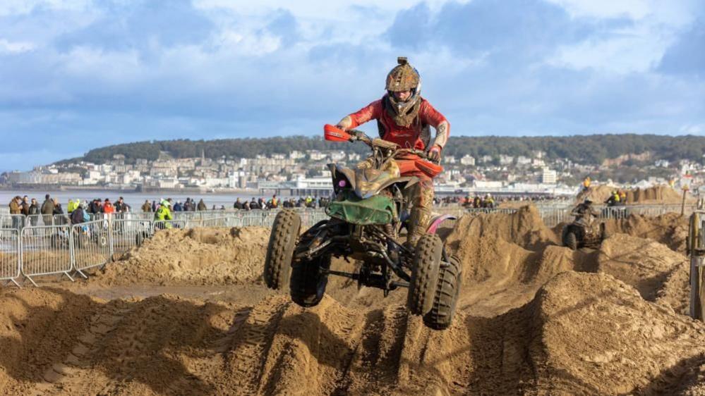 A man riding a quadbike gets significant air while jumping over a mound as part of the Weston Beach Race. Crowds of people and the Weston seafront can be seen in the background, under a mix of cloud and sunny skies.
