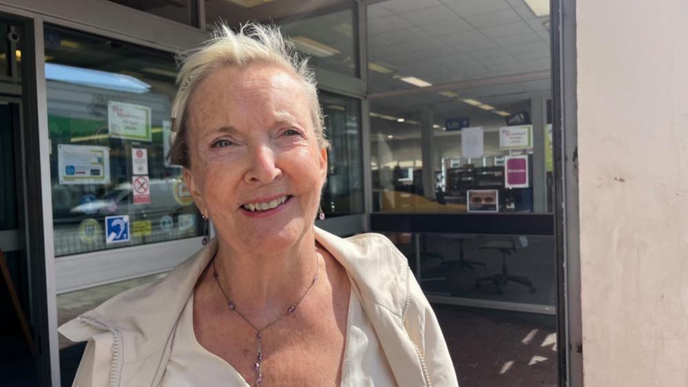 Janet Tough is smiling, by the entrance of Gorleston Library. She has blonde hair, tied back, and is wearing a cream coloured top and jacket. Behind her are the aluminium-framed doors and windows.