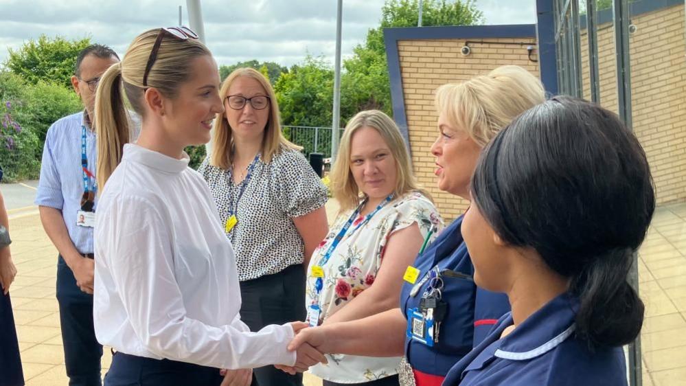 Rosie Wrighting with long blond hair and white top meets two nurses in dark blue uniforms and other staff wearing lanyards outside the hospital