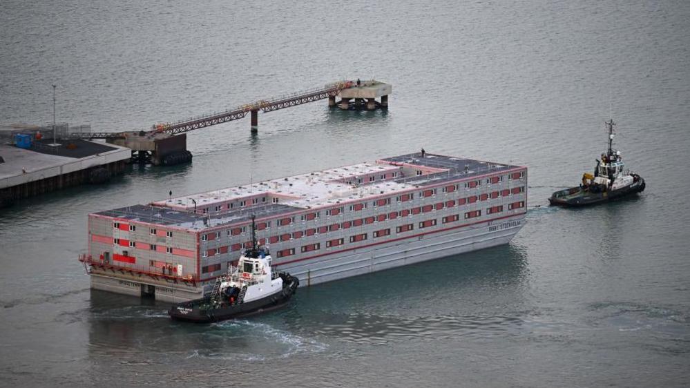 Tugboats tow the Bibby Stockholm out of Portland. The barge is a huge rectangular red and grey boxy vessel, with windows along its sides.