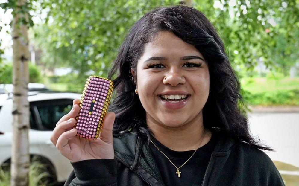 Grace, wearing a black top and jumper, smiles as she holds up her brick-phone, which she has decorated in pink and yellow plastic gems