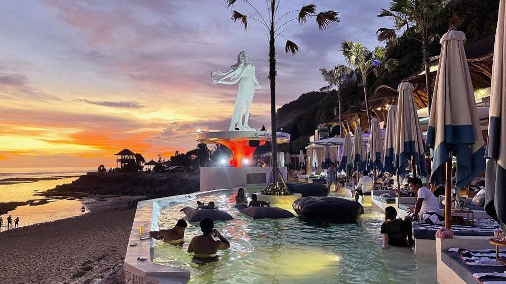 Sunset in the background of a beach, as people relax in a pool at a beach club. There are sunbeds and floaties at the beach club.