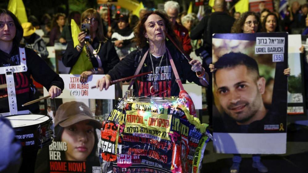 A woman beats a drum as she marches with others who hold flags and placards bearing the faces of hostages