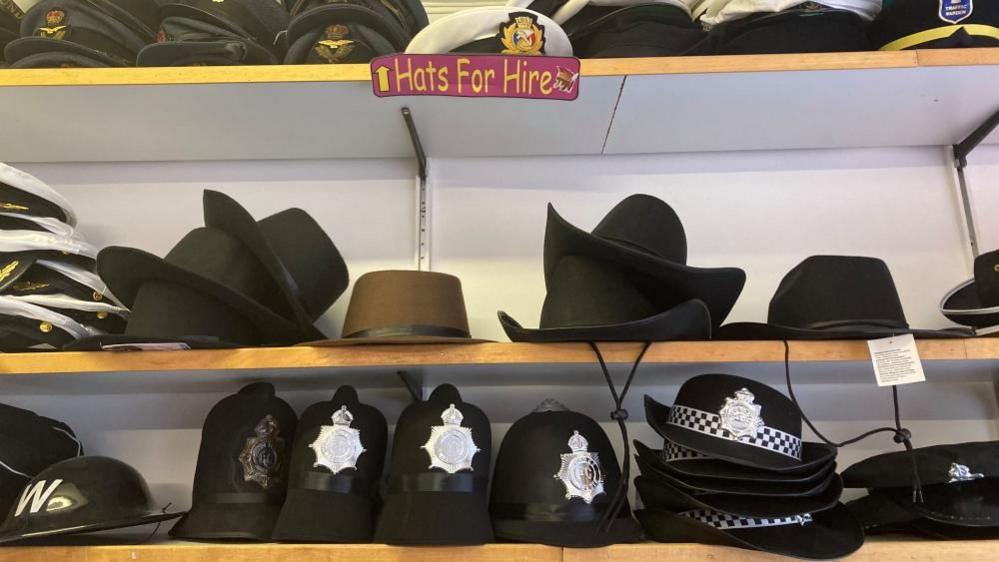 A selection of hats, including police helmets and military caps, sitting on shelves below a sign saying, "Hats For Hire"