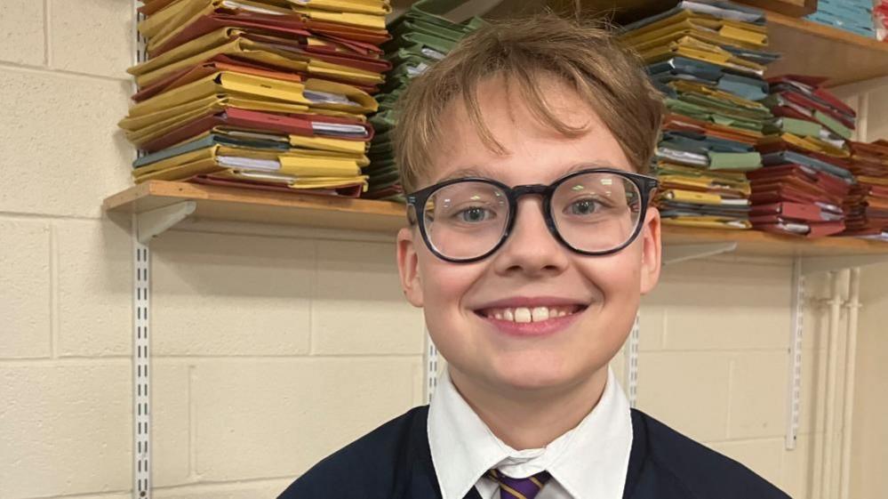 Rojus, standing in front of a shelf of folders. He is wearing glasses, and has brown hair, and is smiling at the camera.