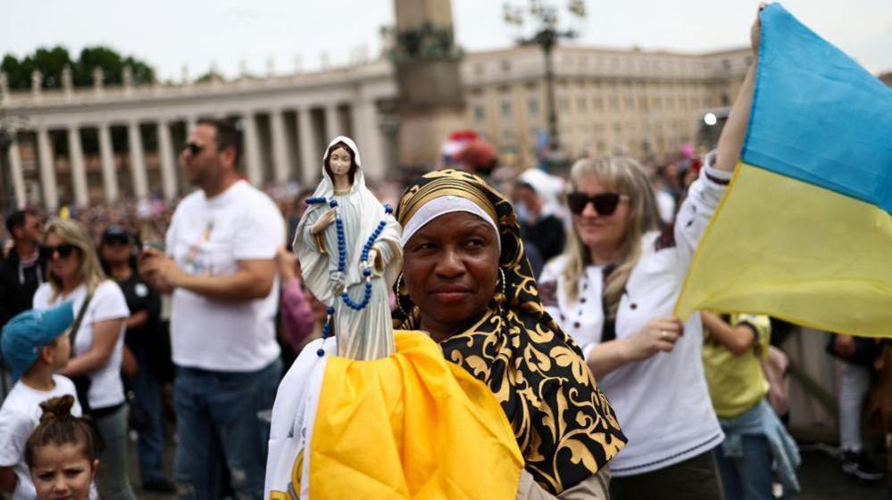 A woman in a vast crowd holds a small statue of the Virgin Mary, while behind her another woman holds a Ukraine flag