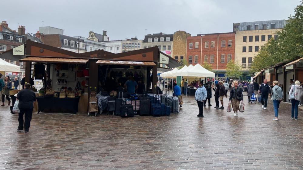 Central fixed brown stall is visible alongside cream-coloured gazebos. Some shoppers are walking by while others are examining the produce.